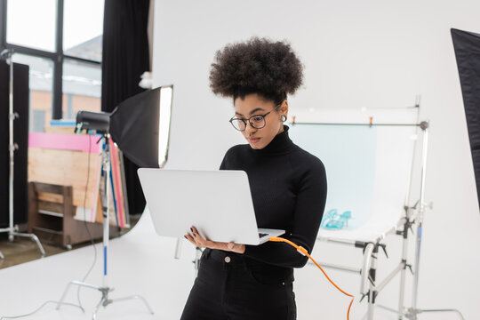 african american content producer in eyeglasses looking at laptop near softbox reflector and shooting table in photo studio.