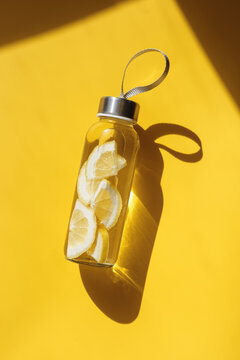 Glass bottle with lemon water drink detox at sunlight on yellow background. Healthy infused water boost metabolism and weight loss. Top view Aesthetic still life, refreshing summer drink