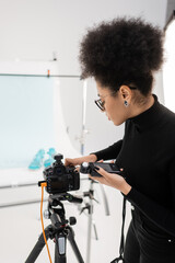 side view of african american content producer with exposure meter adjusting modern digital camera in photo studio.
