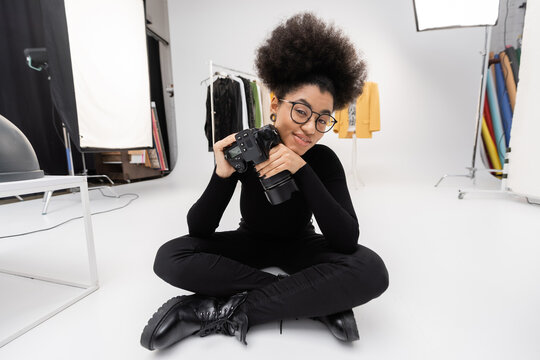smiling african american woman in black clothes sitting with professional digital camera in modern photo studio.