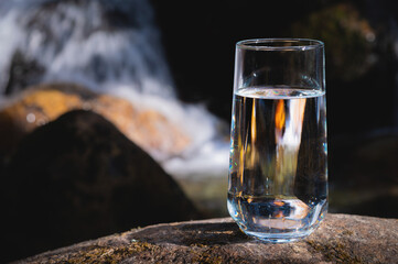 drinking transparent water in a glass glass stands on a stone outdoors in a forest in nature, a waterfall rages on a blurred background