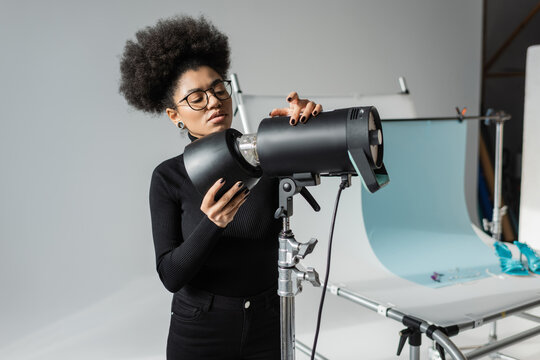 african american content manager in eyeglasses and black turtleneck working with strobe lamp near shooting table in photo studio.