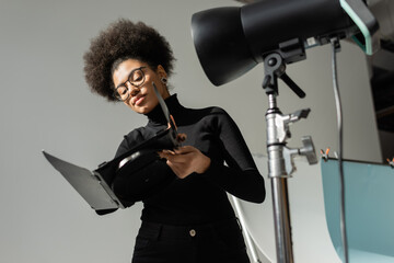 african american content producer in eyeglasses holding part of strobe lamp in photo studio.