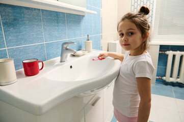 Adorable Caucasian child, lovely little girl in white t-shirt, holding toothbrush, looking at...