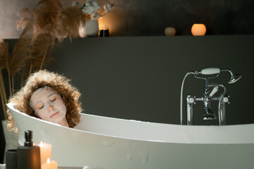 A beautiful woman with curly hair lies in a warm bath with a moisturizing mask on her face. A young woman takes a bath with essential oils.