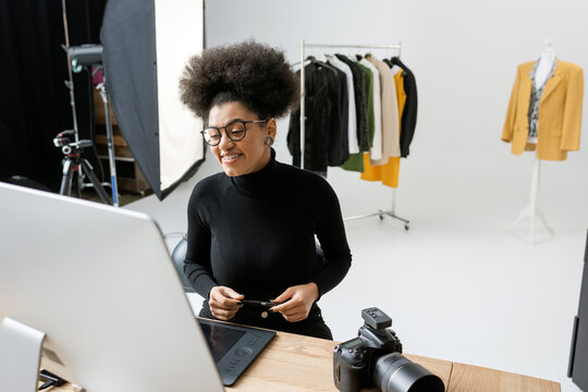 happy african american content maker looking at computer monitor near digital camera and trendy clothes on background in photo studio.