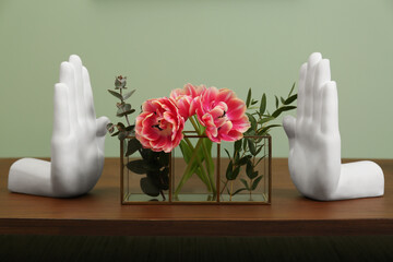 Glass holder with green branches and beautiful flowers near hand shaped bookends on wooden table indoors. Stylish interior design