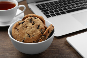 Chocolate chip cookies, cup of tea and laptop on wooden table, closeup. Space for text