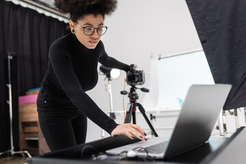 Fototapeta na wymiar african american content producer in black turtleneck and eyeglasses looking at laptop while working in photo studio.