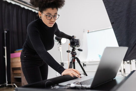 african american content producer in black turtleneck and eyeglasses looking at laptop while working in photo studio.