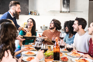Happy multiethnic friends eating spaghetti or noodles with tomato sauce and drinking red wine at home during a amazing dinner party - Smiling hipster guy serving pasta to his friends for dinner
