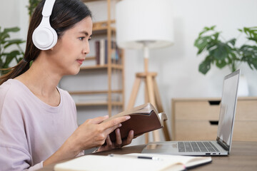 Virtual Lecture. Portrait of young lady in wireless headphones sitting at desk, holding and reading book, having online class with tutor and looking at laptop computer. Woman studying at home