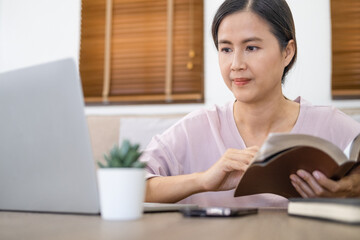 Focused young asian businesswoman or student looking at laptop holding book learning, serious woman working or studying with computer doing research or preparing for exam online.