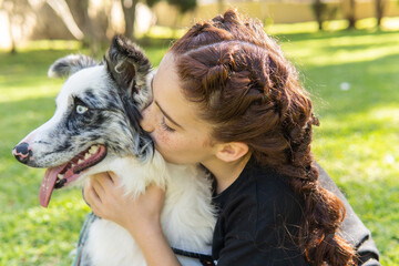  Red-haired girl with pigtails kissing her black and white dog on the grass in a park.