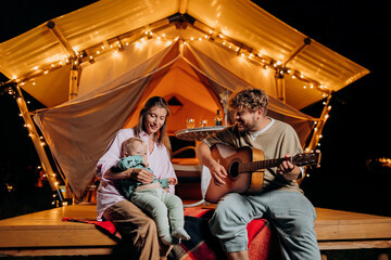 Happy family relaxing and spend time together in glamping on summer evening and playing guitar near cozy bonfire. Luxury camping tent for outdoor recreation and recreation. Lifestyle concept