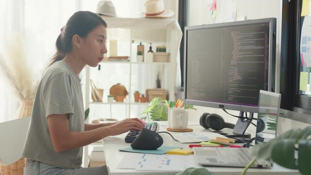 Side view of professional young Asia girl IT development programmer typing on keyboard coding programming fixing data code on computer screen and laptop on table in workroom at house office.