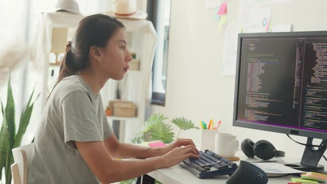 Side view of professional young Asia girl IT development programmer typing on keyboard coding programming fixing data code on computer screen and laptop on table in workroom at house office.