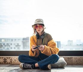 Copy space. Beautiful teenage girl, internet influencer sitting on patio floor on balcony, using smartphone, taking a selfie and entertainment. Yellow fur coat and red glasses. City in background.