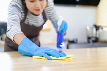 Asian housewife sanitizing surfaces cleaning home kitchen table with disinfectant spray bottle washing surface with towel and gloves.