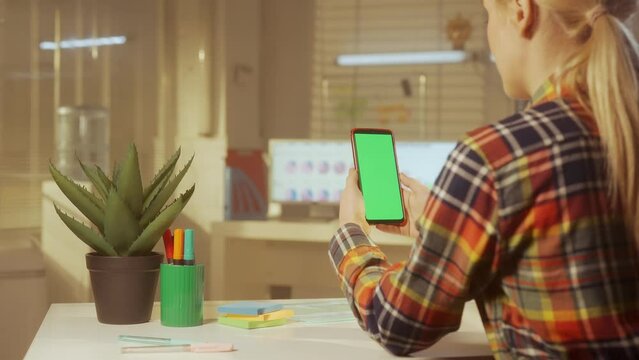 Back view of a young woman is holding a mobile phone with a green screen. A woman manager, designer or programmer in a colorful checkered shirt. Office work. Advertising area, workspace mock up.