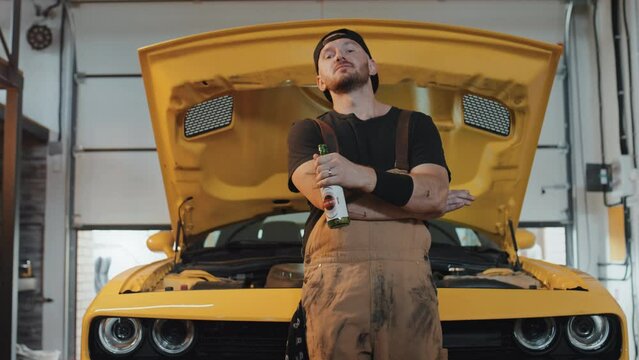 Medium low angle shot of mechanic wearing coveralls and black cap backwards standing in front of yellow opened motor hood, holding beer, crossing his arms, then looking at camera. Parking house