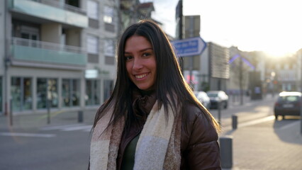 Obraz premium Portrait of a beautiful young woman standing in city street during sunset time with sunlight flare shining. Happy expression of a brunette adult girl in 20s smiling at camera