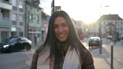 Obraz premium Portrait of a beautiful young woman standing in city street during sunset time with sunlight flare shining. Happy expression of a brunette adult girl in 20s smiling at camera