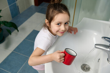 Mischievous baby girl standing by sink in home bathroom, holding red cup of water, smiling looking...
