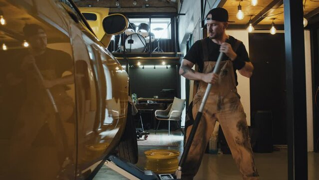Medium full shot of tattooed auto mechanic wearing coveralls and black cap backwards moving ram lever, lifting body of yellow car. Modern parking house, daytime