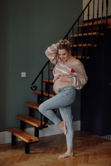 Portrait of young pregnant woman wear beige sweater with application of heart, jeans, raising leg, showing naked belly.
