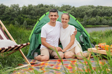 Naklejka na ściany i meble Smiling happy optimistic woman and man wearing casual clothing sitting at tent near the river looking at camera having picnic enjoying spring days.