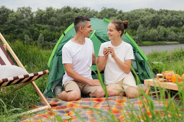 Obraz na płótnie Canvas Vacation in nature. Loving woman and man wearing casual clothing sitting at tent near the river, enjoyoing hot coffee or tea, looking at each other, smiling happily.