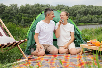 Tasty coffee on nature. Joyful smiling woman and man wearing casual clothing sitting at tent near the river, drinking coffee, enjoying aromat, resting, relaxing.