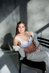 A pregnant pretty woman in a white suit and bra is sitting on a gray background. Pregnant cute woman stroking her belly and smiling. Happy period of pregnancy