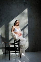 Fototapeta na wymiar A beautiful woman with fair hair is pregnant in a white elegant suit sitting on a chair on a gray background. Fashion for pregnant women. A stylish woman is pregnant.