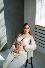 A beautiful woman with fair hair is pregnant in a white elegant suit sitting on a chair on a gray background. Fashion for pregnant women. A stylish woman is pregnant.