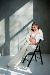 A beautiful woman with fair hair is pregnant in a white elegant suit sitting on a chair on a gray background. Fashion for pregnant women. A stylish woman is pregnant.