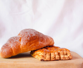 Dos panes dulces, uno sobre otro en fondo blanco sobre una tabla de madera, un cuernito o croissant con azúcar y un pan de miel