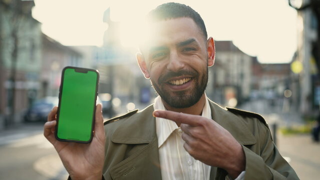 One cheerful Arab guy pointing at phone with greenscreen in chroma key standing in urban street. A Middle Eastern male person pointing at online content display in cellphone modern tech device