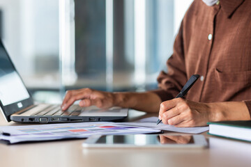 Close-up photo. Hands of a young woman in a brown shirt of a student writing with a pen, studying online at a laptop.