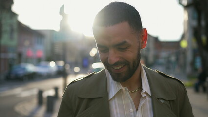 One happy handsome Moroccan young man smiling standing in city street with afternoon lens flare sunlight. Candid and authentic lifestyle