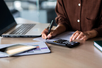 Close-up photo. Hands of a young woman in a brown shirt calculating on a calculator and writing documents at an office table.