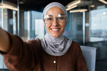  Smiling businesswoman in hijab standing in office and talking on video call from phone. Explains, consults, business meeting.