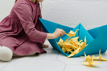  A little girl is playing holding a yellow origami crane in her hand and putting it in a blue ship