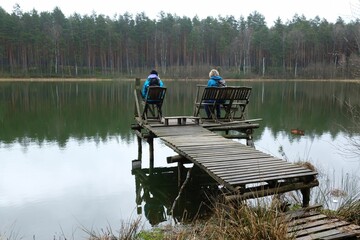 Fototapeta na wymiar Two women in blue jackets are sitting on a wooden pier by the river on a spring day.