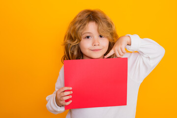 Child showing index finger on red sheet of paper, isolated on yellow background. Portrait of a kid holding a blank placard, poster. Kind kids face, amazed emotions of child.