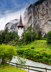 Fototapeta na wymiar Lauterbrunnen church with the Staubbach falls cascading down into the valley from the cliffside of the Swiss Alps on a summer morning with low cloud cover