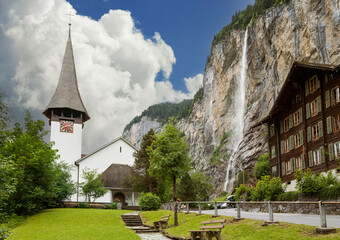 Lauterbrunnen church with the Staubbach falls cascading down into the valley from the cliffside of the Swiss Alps on a summer morning with low cloud cover