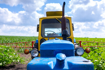 An old farm tractor painted in yellow-blue color with a trailed sprayer with tank capacities is...
