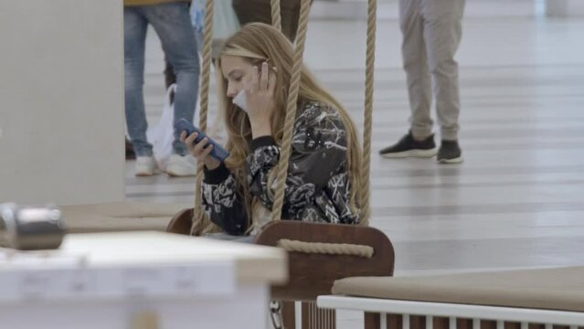 Young girl in medical mask using smartphone while swinging in a shopping center. Stock footage. Public places during coronavirus.